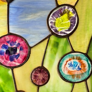 K&ouml;nnte beinhalten: Ein Buntglasfenster mit einem Netzwerk aus dunklen Metalllinien, die verschiedene kreisf&ouml;rmige Glaskabochons in Violett-, Rosa-, Blau- und Oranget&ouml;nen verbinden, vor einem Hintergrund aus strukturierten gelben, blauen und gr&uuml;nen Glasscheiben.