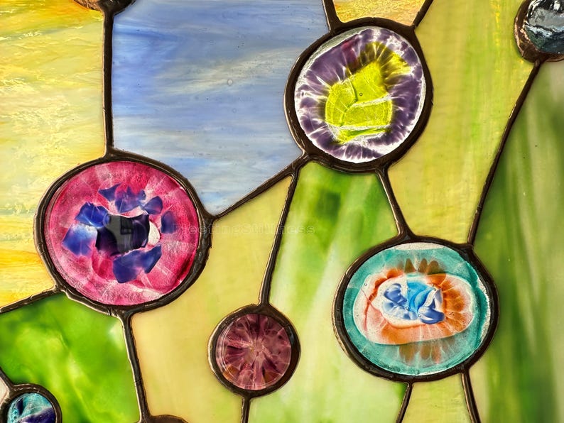 K&ouml;nnte beinhalten: Ein Buntglasfenster mit einem Netzwerk aus dunklen Metalllinien, die verschiedene kreisf&ouml;rmige Glaskabochons in Violett-, Rosa-, Blau- und Oranget&ouml;nen verbinden, vor einem Hintergrund aus strukturierten gelben, blauen und gr&uuml;nen Glasscheiben.