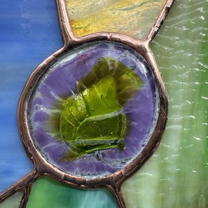 K&ouml;nnte beinhalten: Ein Buntglasfenster mit einem zentralen, kreisf&ouml;rmigen St&uuml;ck aus texturiertem lindgr&uuml;nem Glas, das von einem violetten Rand umgeben und mit Kupferfolien-L&ouml;tlinien eingefasst ist. Die umliegenden Glasscheiben sind blau, gelb und hellgr&uuml;n.