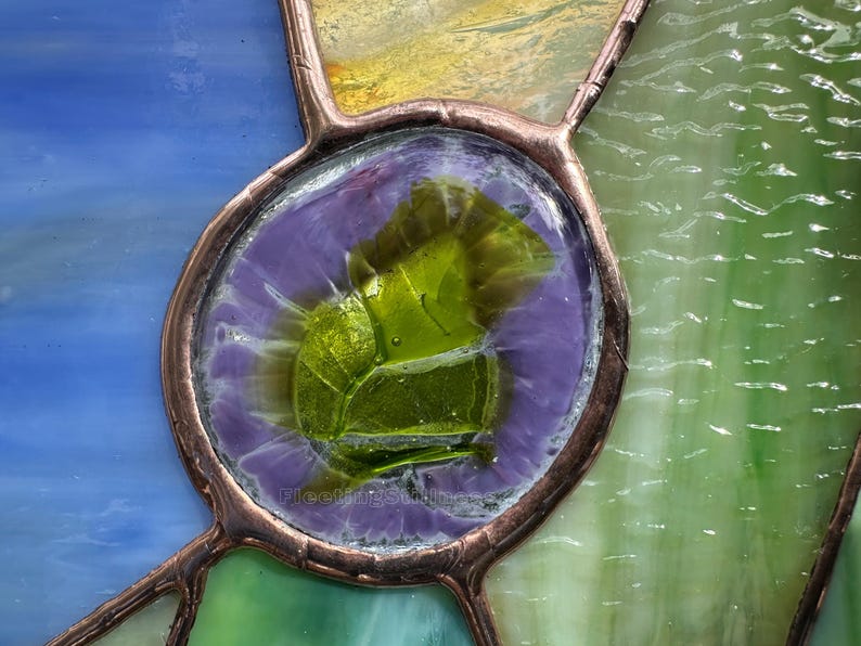 K&ouml;nnte beinhalten: Ein Buntglasfenster mit einem zentralen, kreisf&ouml;rmigen St&uuml;ck aus texturiertem lindgr&uuml;nem Glas, das von einem violetten Rand umgeben und mit Kupferfolien-L&ouml;tlinien eingefasst ist. Die umliegenden Glasscheiben sind blau, gelb und hellgr&uuml;n.