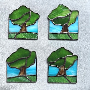 May include: A set of four square stained glass coasters, each featuring a landscape design with a brown tree trunk, green glass foliage, a light blue sky, and a textured green glass foreground, all held together by dark metal solder lines.