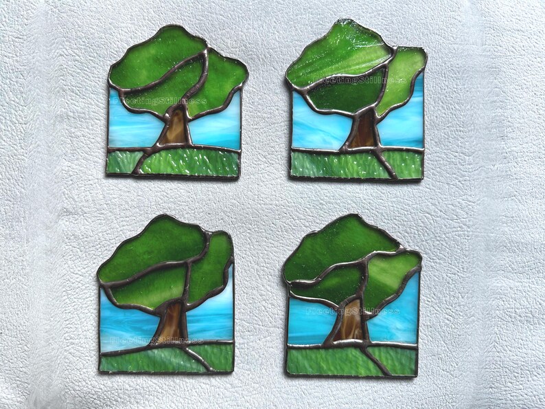May include: A set of four square stained glass coasters, each featuring a landscape design with a brown tree trunk, green glass foliage, a light blue sky, and a textured green glass foreground, all held together by dark metal solder lines.