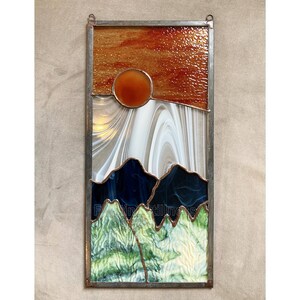 May include: A stained glass window depicting a sunset over mountains. The sky is a gradient of orange, red, and yellow. The mountains are a dark blue and green. The sun is a bright orange circle.