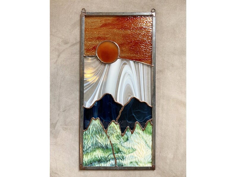 May include: A stained glass window depicting a sunset over mountains. The sky is a gradient of orange, red, and yellow. The mountains are a dark blue and green. The sun is a bright orange circle.
