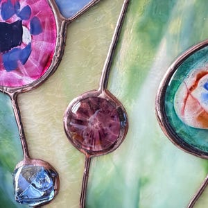 K&ouml;nnte beinhalten: Ein Buntglasfenster mit einem Kupferfolien-L&ouml;t-Design und verschiedenen Glaselementen. Es enth&auml;lt ein gro&szlig;es kreisf&ouml;rmiges St&uuml;ck in Pink und Blau, einen kleineren runden violetten Akzent und eine ovale Form in Blaugr&uuml;n, Blau und Orange.