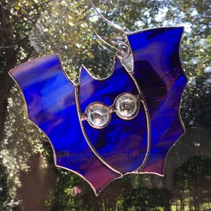 May include: A stained glass bat ornament with blue and purple glass and clear crystal eyes. The bat is hanging on a window.