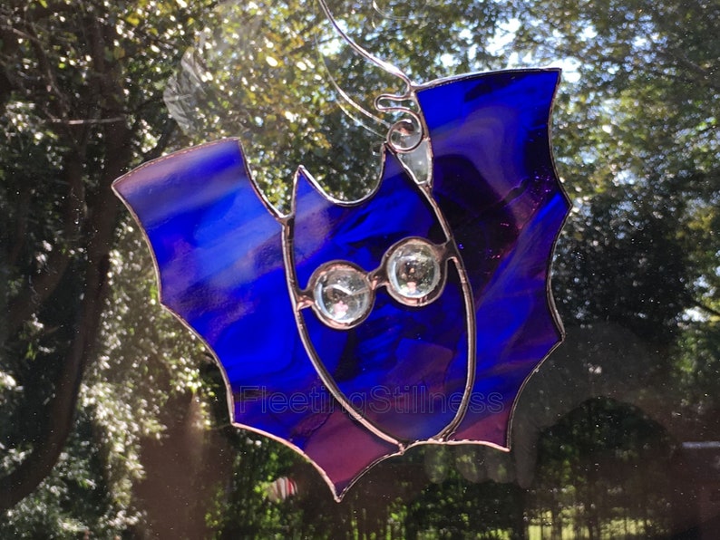 May include: A stained glass bat ornament with blue and purple glass and clear crystal eyes. The bat is hanging on a window.