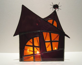 Könnte beinhalten: Ein Halloween-Haus aus Buntglas in dunklem Lila. Das Haus hat ein spitzes Dach und gewölbte Fenster mit einer kleinen Spinne oben drauf. Die Fenster leuchten orange, was auf Kerzen im Inneren hindeutet. Ein dekoratives Stück für die gruselige Jahreszeit.