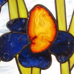 May include: Close-up of a stained glass window featuring a blue and yellow flower with an orange center. The center of the flower is made of a translucent orange material with a swirling pattern.