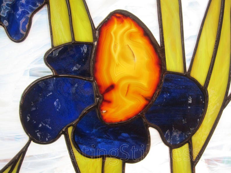 May include: Close-up of a stained glass window featuring a blue and yellow flower with an orange center. The center of the flower is made of a translucent orange material with a swirling pattern.