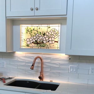 May include: A rectangular stained glass panel featuring white dogwood flowers with yellow centers and green foliage, set into a white kitchen cabinet frame above a copper faucet and a black double sink with white subway tile backsplash.