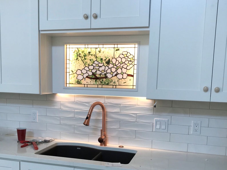 May include: A rectangular stained glass panel featuring white dogwood flowers with yellow centers and green foliage, set into a white kitchen cabinet frame above a copper faucet and a black double sink with white subway tile backsplash.