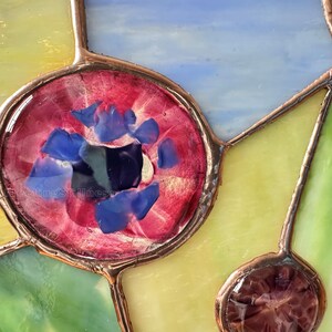 K&ouml;nnte beinhalten: Ein Buntglasfenster mit einem kreisf&ouml;rmigen roten Glaselement, das mit kleinen blauen Glassplittern gef&uuml;llt ist. Das St&uuml;ck ist mit Kupferfolien-L&ouml;tlinien konstruiert, die verschiedene durchscheinende Glassegmente in Gelb, Gr&uuml;n und Hellblau verbinden.