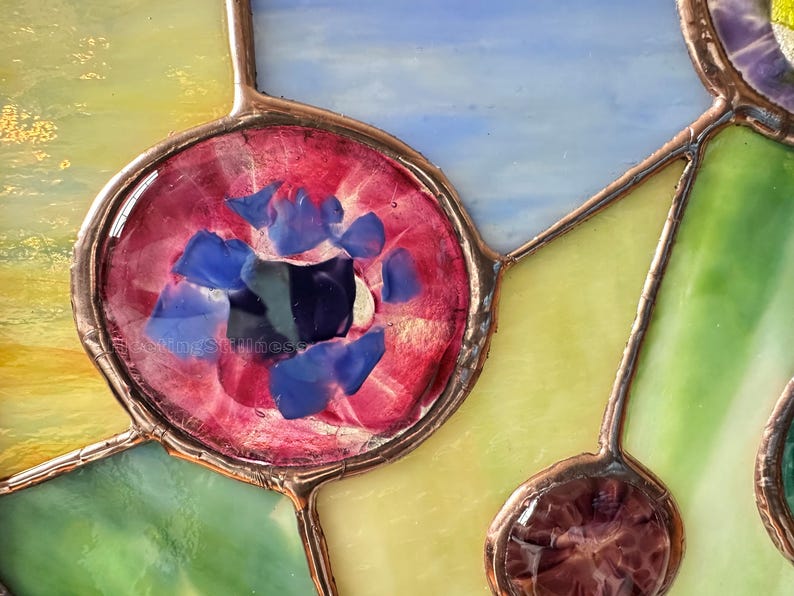 K&ouml;nnte beinhalten: Ein Buntglasfenster mit einem kreisf&ouml;rmigen roten Glaselement, das mit kleinen blauen Glassplittern gef&uuml;llt ist. Das St&uuml;ck ist mit Kupferfolien-L&ouml;tlinien konstruiert, die verschiedene durchscheinende Glassegmente in Gelb, Gr&uuml;n und Hellblau verbinden.