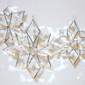 May include: Three glass snowflake ornaments with a silver finish and gold accents. The snowflakes are arranged in a row, with the largest snowflake in the center.