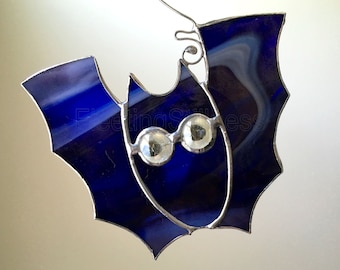 Könnte beinhalten: Eine Fledermaus aus Buntglas mit blauem und weißem Glas und silbernen Glitzeraugen. Die Fledermaus hängt an einem silbernen Draht.