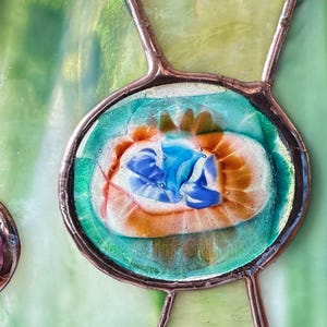 K&ouml;nnte beinhalten: Ein Buntglasfenster mit einem zentralen ovalen Element aus blauem, wei&szlig;em und orangefarbenem Wirbelglas, das von einer Kupferfolie eingefasst ist. Das umgebende Glas ist hellgr&uuml;n mit strukturierter Oberfl&auml;che und durch d&uuml;nne Kupferl&ouml;tstellen verbunden.