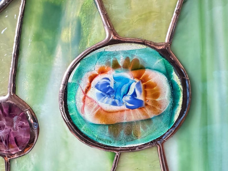 K&ouml;nnte beinhalten: Ein Buntglasfenster mit einem zentralen ovalen Element aus blauem, wei&szlig;em und orangefarbenem Wirbelglas, das von einer Kupferfolie eingefasst ist. Das umgebende Glas ist hellgr&uuml;n mit strukturierter Oberfl&auml;che und durch d&uuml;nne Kupferl&ouml;tstellen verbunden.