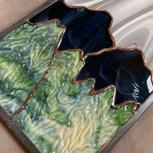 May include: A stained glass artwork depicting a mountain range with green, blue, and white glass pieces. The glass is framed in a dark metal frame.