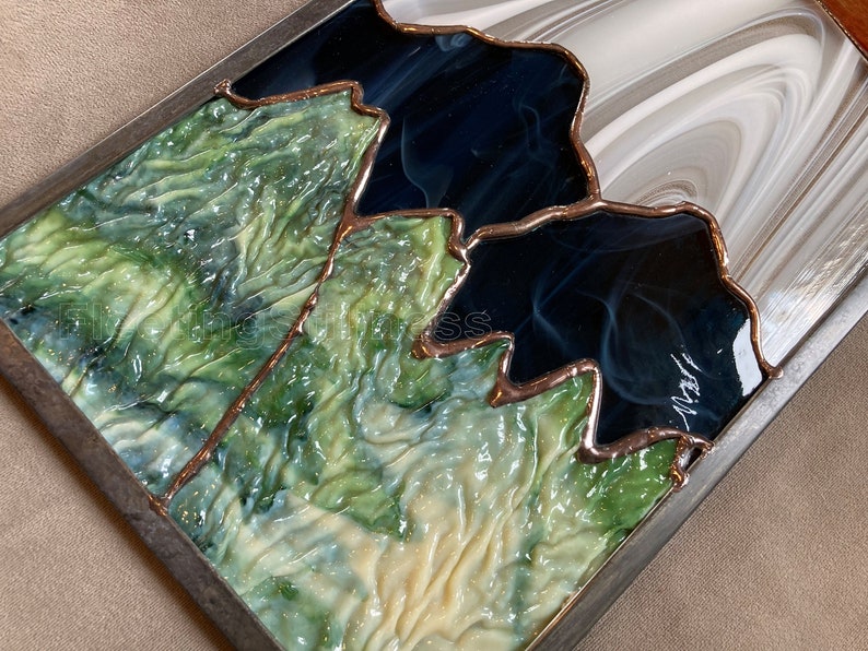 May include: A stained glass artwork depicting a mountain range with green, blue, and white glass pieces. The glass is framed in a dark metal frame.