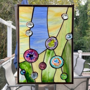 K&ouml;nnte beinhalten: Ein rechteckiges Buntglasfenster mit einer geometrischen Komposition aus gelben, blauen und gr&uuml;nen Glassegmenten. Das St&uuml;ck ist mit verschiedenen kreisf&ouml;rmigen Glas-Cabochons in Rosa, Lila, T&uuml;rkis und Transparent verziert, die durch dunkle Metalll&ouml;tlinien zusammengehalten werden.