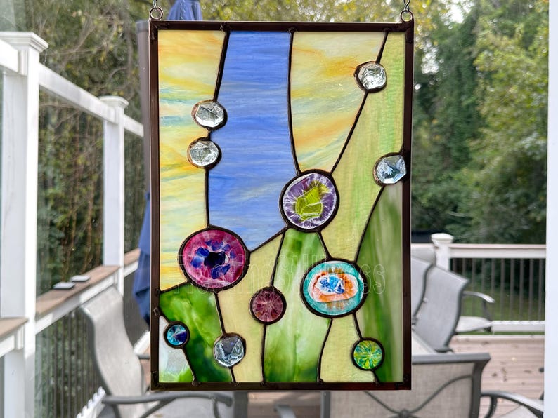 K&ouml;nnte beinhalten: Ein rechteckiges Buntglasfenster mit einer geometrischen Komposition aus gelben, blauen und gr&uuml;nen Glassegmenten. Das St&uuml;ck ist mit verschiedenen kreisf&ouml;rmigen Glas-Cabochons in Rosa, Lila, T&uuml;rkis und Transparent verziert, die durch dunkle Metalll&ouml;tlinien zusammengehalten werden.