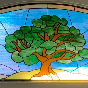 May include: A stained glass window depicting a green tree with brown branches against a blue sky with white clouds. The tree is in the center of the window and is surrounded by a light brown landscape.