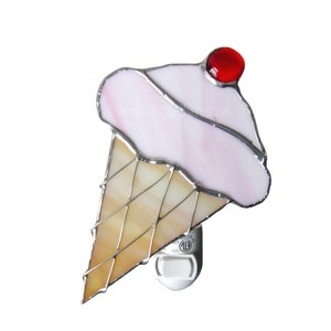 May include: A stained glass ice cream cone night light with a pink ice cream scoop and a red cherry on top. The cone is a light brown color and the night light is plugged into a standard electrical outlet.