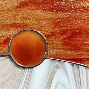 May include: A stained glass panel with a circular orange piece of glass in the center. The panel is set in a white frame and has a brown and red background.