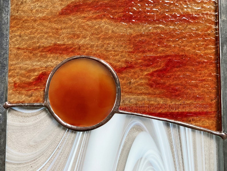 May include: A stained glass panel with a circular orange piece of glass in the center. The panel is set in a white frame and has a brown and red background.