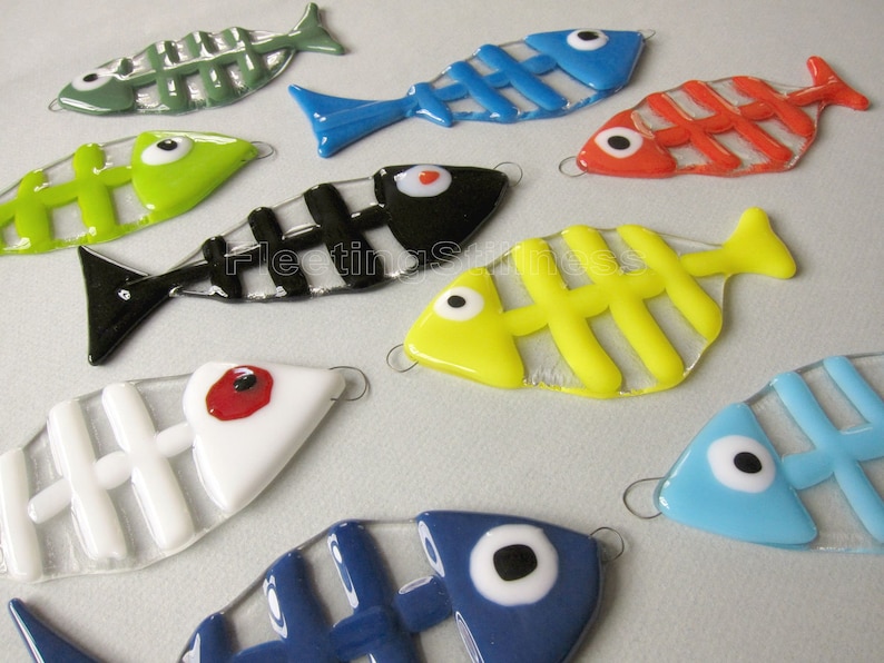 K&ouml;nnte beinhalten: Eine Sammlung von neun Glasfisch-Ornamenten mit bunten Skeletten und wei&szlig;en Augen. Die Fische sind in verschiedenen Farben, darunter Gr&uuml;n, Rot, Blau, Gelb, Schwarz und Wei&szlig;.