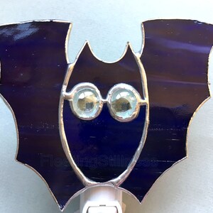 May include: A stained glass bat nightlight with iridescent blue wings and clear glass eyes. The nightlight is plugged into an outlet.
