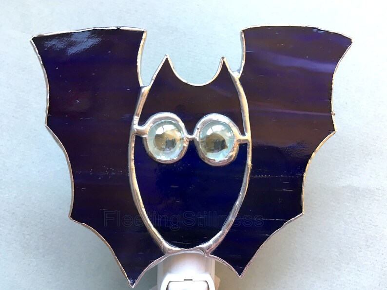 May include: A stained glass bat nightlight with iridescent blue wings and clear glass eyes. The nightlight is plugged into an outlet.