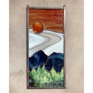 May include: A stained glass window depicting a sunset over a mountain range. The sun is a bright orange circle, and the mountains are a mix of blue, green, and white glass.