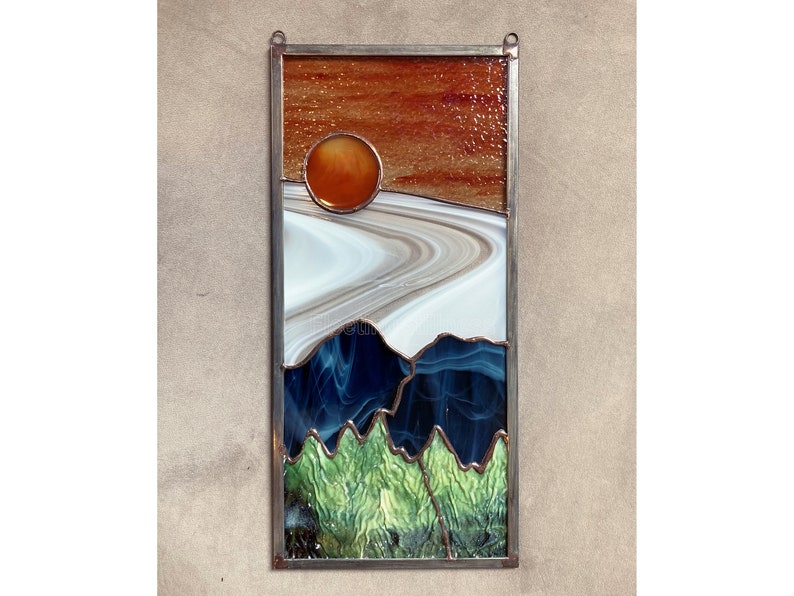 May include: A stained glass window depicting a sunset over a mountain range. The sun is a bright orange circle, and the mountains are a mix of blue, green, and white glass.