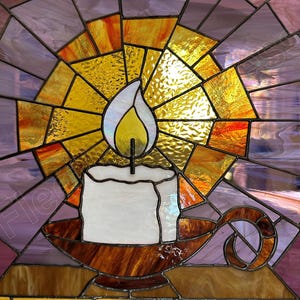K&ouml;nnte beinhalten: Buntglasfenster mit einer brennenden Kerze in einem Halter. Die Kerze ist wei&szlig; mit einer gelben Flamme, vor einem Hintergrund aus strahlenden gelben, orangefarbenen und violetten Glasteilen. Der Kerzenhalter ist braun.