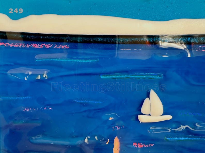 May include: A fused glass art piece featuring a deep blue ocean scene with a white sailboat, a white shoreline, and the text 249 and FleetingStillness. The composition includes horizontal blue glass layers, small white accents, and pinkish details.