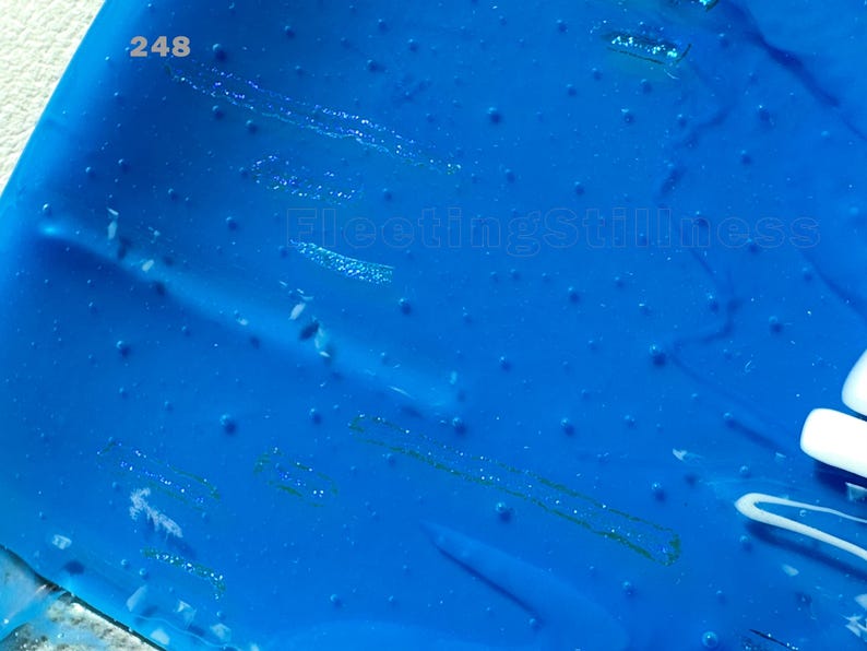 May include: A vibrant blue translucent surface featuring a scattered pattern of small raised dots and subtle linear indentations. The number 248 is printed in the upper left corner, and the text FleetingStillness appears in the center.