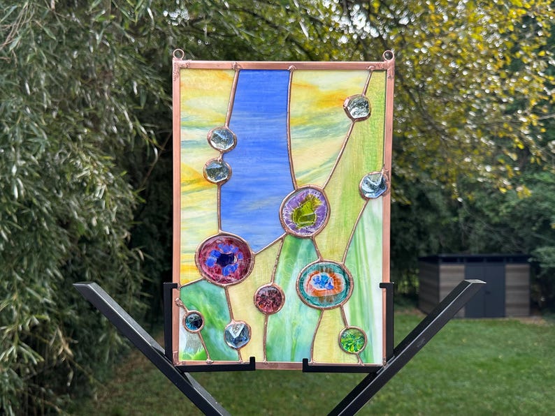 May include: A rectangular stained glass panel framed in copper, featuring a vertical composition of yellow, blue, and green glass segments accented by various small, circular, and multi-colored glass cabochons soldered onto the surface.
