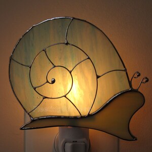 May include: A stained glass night light shaped like a snail with a yellow shell and a green spiral. The light is plugged into an outlet and is lit.
