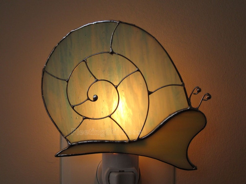 May include: A stained glass night light shaped like a snail with a yellow shell and a green spiral. The light is plugged into an outlet and is lit.