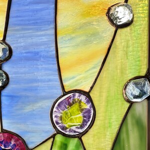 K&ouml;nnte beinhalten: Ein Buntglasfenster mit strukturierten gelben, blauen und gr&uuml;nen Glassegmenten, die durch dunkle Metalll&ouml;tlinien verbunden sind. Das Design enth&auml;lt mehrere klare, facettierte Glasschmucksteine sowie zwei runde Glaskabochons in Violett und Pink.