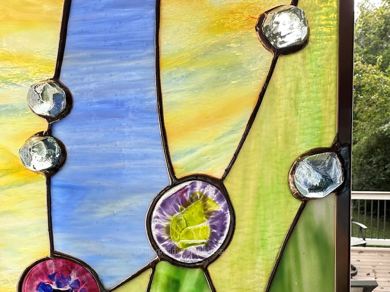 K&ouml;nnte beinhalten: Ein Buntglasfenster mit strukturierten gelben, blauen und gr&uuml;nen Glassegmenten, die durch dunkle Metalll&ouml;tlinien verbunden sind. Das Design enth&auml;lt mehrere klare, facettierte Glasschmucksteine sowie zwei runde Glaskabochons in Violett und Pink.