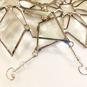 May include: A silver-toned, geometric, stained glass snowflake ornament with a clear glass center and a beaded hanging loop.