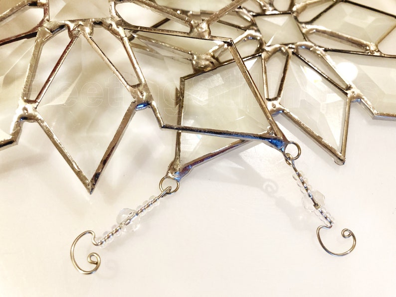 May include: A silver-toned, geometric, stained glass snowflake ornament with a clear glass center and a beaded hanging loop.