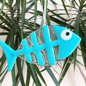 K&ouml;nnte beinhalten: Ein t&uuml;rkisblauer Glasfisch-Anh&auml;nger mit einem wei&szlig;en Auge und einer schwarzen Pupille. Der Fisch hat einen klaren Glask&ouml;rper mit einem t&uuml;rkisblauen Skelettmuster.