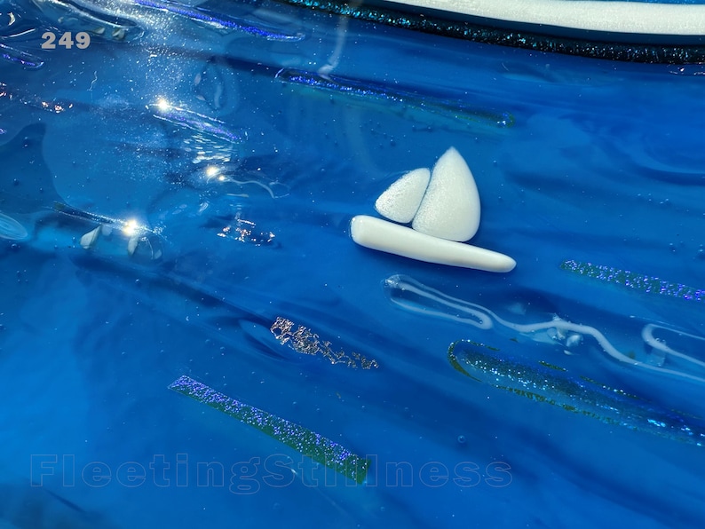May include: A glossy blue glass surface featuring a small white sailboat icon, iridescent glitter accents, and the text 249 in the upper left corner and FleetingStillness at the bottom. The surface has a wavy, fluid texture with light reflections.