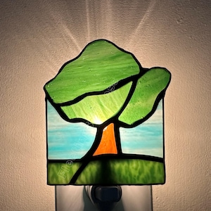 May include: A stained glass night light featuring a tree design with a green canopy, an orange trunk, and a light blue sky background. The rectangular piece is illuminated from behind, casting a soft glow against the wall behind the fixture.