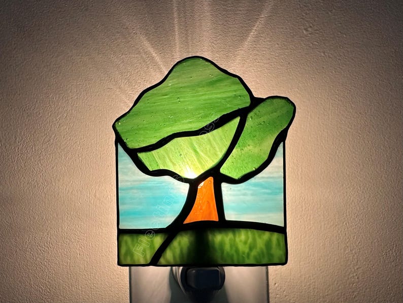 May include: A stained glass night light featuring a tree design with a green canopy, an orange trunk, and a light blue sky background. The rectangular piece is illuminated from behind, casting a soft glow against the wall behind the fixture.