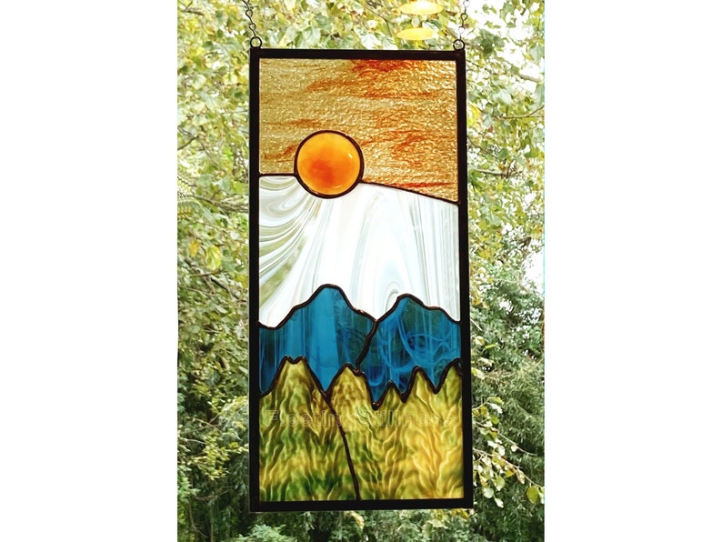 May include: A stained glass window depicting a sunset over a mountain range. The sun is a bright orange circle, and the mountains are a mix of blue and green.